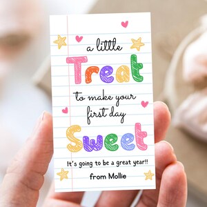 May include: A rectangular gift tag with a white lined paper background featuring colorful scribbled text that reads: a little Treat to make your first day Sweet. It's going to be a great year!! from Mollie, decorated with small pink hearts and yellow stars.