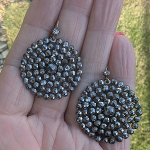 May include: A pair of dark grey, circular earrings. Each earring features many small, faceted beads arranged in a circular design. A small, clear gem is at the top of each earring. The earrings are held in a person's hand.