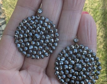 May include: A pair of dark gray, round earrings. Each earring is composed of numerous small, faceted beads arranged in a circular pattern. A small, clear gem is at the top of each earring. The earrings are held in a person's hand.
