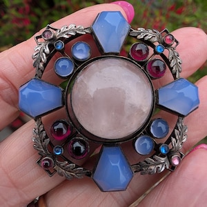 May include: A circular brooch featuring a large central pale pink cabochon stone surrounded by four faceted light blue stones in a coffin shape. The metal frame is decorated with leaf motifs and accented with smaller round blue and deep red stones.