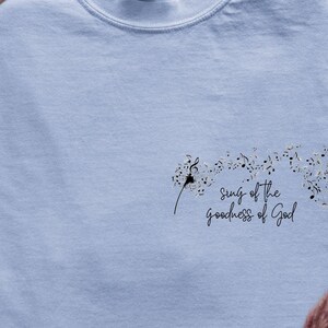 Könnte beinhalten: Ein hellblaues T-Shirt mit einem Löwenzahnmuster und dem Text "Sing of the goodness of God".