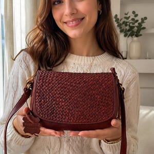 May include: A dark red, woven leather crossbody bag with a rounded flap closure and a matching adjustable shoulder strap. A small, leaf-shaped leather tag is attached to the side of the bag, featuring embossed text that reads Genuine Leather.