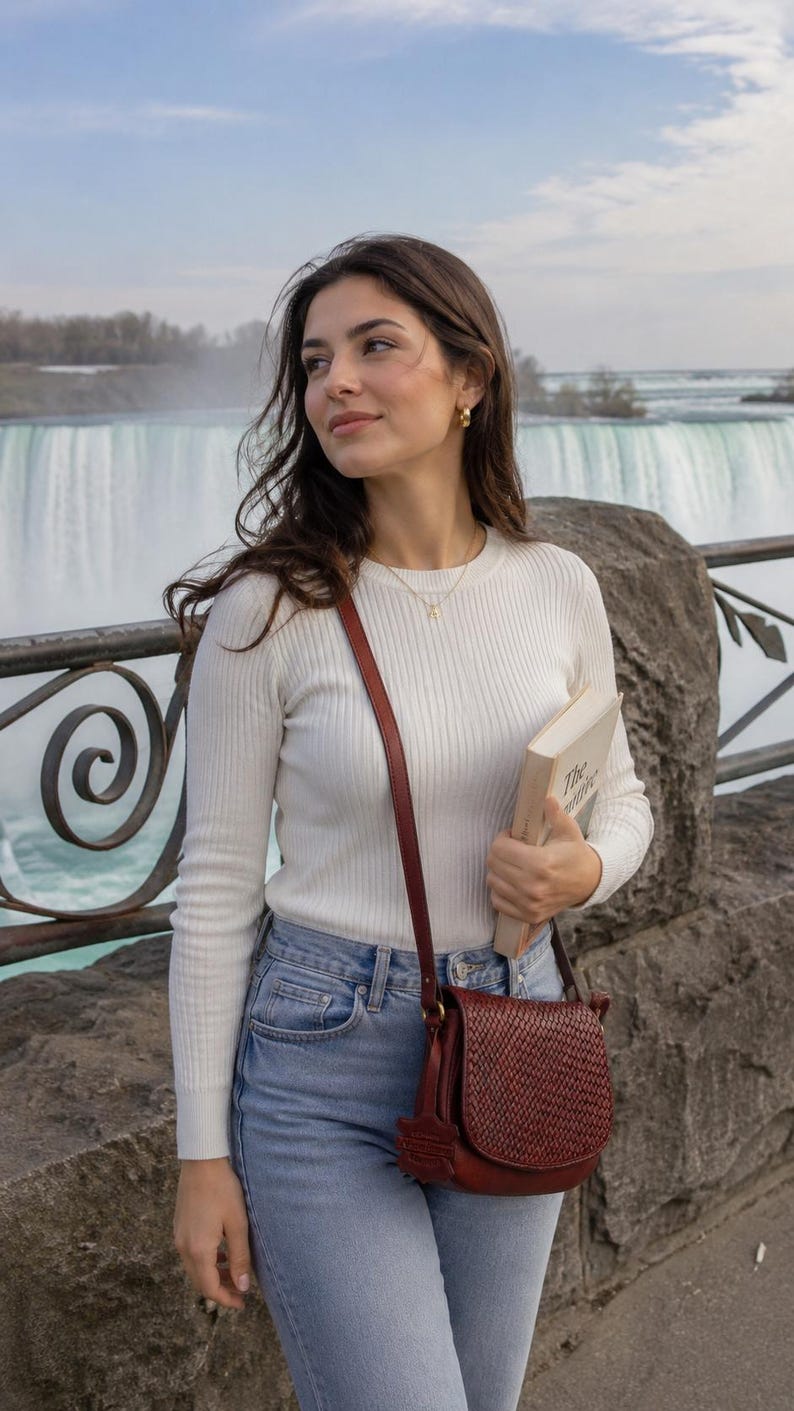 May include: A white long-sleeved ribbed sweater, light blue denim jeans, and a dark red woven leather crossbody bag. The bag features a flap closure and a thin shoulder strap. A book with a cream-colored cover is held in one hand.