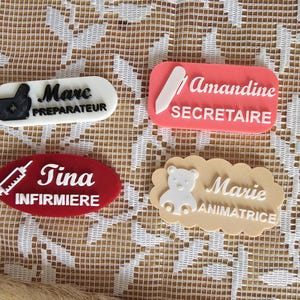 Peut inclure: Quatre badges nominatifs personnalisés avec lettrage en relief et icônes. Un badge ovale blanc indique Marc PREPARATEUR avec un mortier. Un badge rectangulaire rose indique Amandine SECRETAIRE avec un stylo. Un badge ovale rouge indique Tina INFIRMIERE avec une seringue. Un badge beige en forme de nuage indique Marie ANIMATRICE avec un ours.