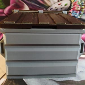 May include: A small, gray plastic toy dumpster with a brown lid. The lid is closed and has a slight curve to it.