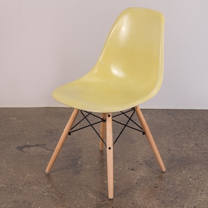 May include: A yellow fiberglass chair with a black metal frame and wooden legs. The chair is on a concrete floor.