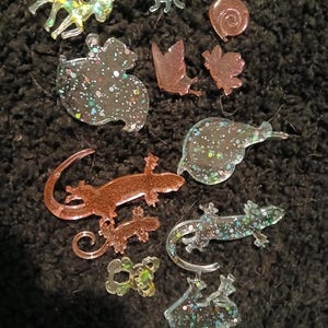 May include: A collection of 10 glitter resin charms in various shapes, including a deer, butterfly, snail, ladybug, gecko, and frog. The charms are translucent and have a sparkly finish.