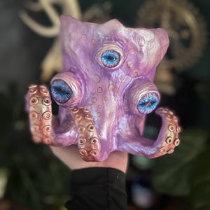Puede incluir: Un macetero de cerámica morado en forma de pulpo con tres ojos grandes azules y detalles dorados. El pulpo tiene ocho tentáculos con ventosas.
