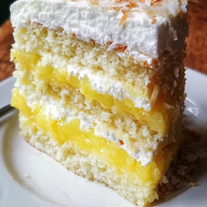 May include: A slice of pineapple coconut cake on a white plate. The cake has layers of yellow pineapple filling, white frosting, and a coconut topping. The text "PINEAPPLE COCONUT CAKE" is at the top of the image.