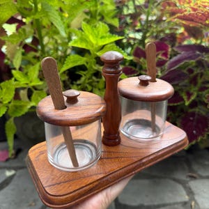 May include: A wooden condiment set featuring two glass jars with wooden lids and spoons, set on a wooden tray. The jars are clear, and the wood has a warm, brown tone. The set is designed for serving spices or condiments.