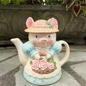 May include: A ceramic teapot shaped like a pig wearing a straw hat with a pink rose. The pig is dressed in a blue-sleeved top and a white apron with a basket of pink roses. The teapot has a spout and handle.