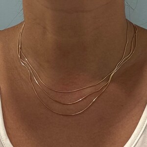 Puede incluir: Un delicado collar de varias hebras en tono dorado que presenta tres cadenas finas y lisas que caen en curvas superpuestas alrededor del cuello. Las cadenas son uniformes en textura y color, creando un aspecto sencillo y superpuesto.