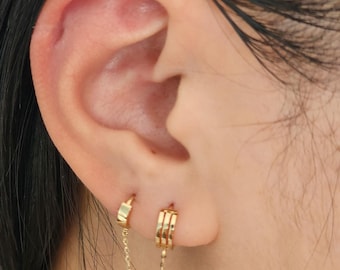 May include: Gold hoop earrings with a delicate chain connecting them. The earrings are worn in the upper lobe of the ear.