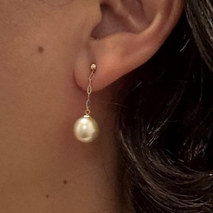 Puede incluir: Primer plano de un pendiente dorado con una perla blanca colgante. El pendiente está sujeto a la oreja con un pequeño perno de oro y una delicada cadena. La perla es redonda y brillante.