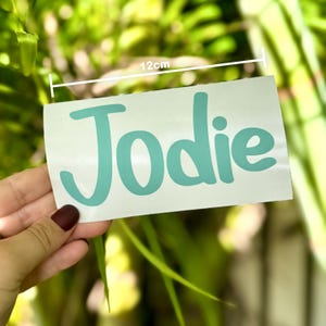 May include: A teal blue vinyl sticker with the name "Jodie" written in a cursive font. The sticker is 12 cm wide.