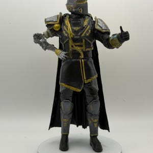 Pode incluir: Uma figura de ação em pé vestindo armadura preta e prateada com detalhes dourados. A figura apresenta um capacete, uma longa capa preta e um braço direito com aparência mecânica. A mão esquerda faz um gesto de polegar para cima em base circular.