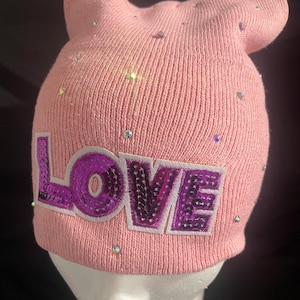 Puede incluir: Un gorro de punto rosa claro con orejas de gato y la palabra "LOVE" en letras de lentejuelas moradas. El gorro está adornado con pedrería iridiscente dispersa. Un accesorio divertido y elegante.