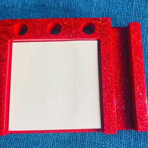 May include: A red glitter desk organiser with three circular holes at the top and a rectangular slot on the side. The organiser has a white notepad in the centre.