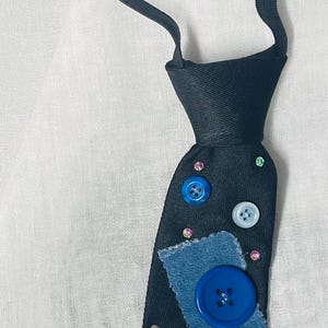 Puede incluir: Una corbata negra con un diseño decorativo. La corbata presenta parches de mezclilla azul, botones de varios tamaños en azul y blanco y pequeños pedrería iridiscente. La corbata tiene una banda elástica negra ajustable.