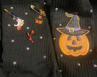 Puede incluir: Calcetines negros con adornos de Halloween. Uno presenta una bruja en una escoba y un alfiler de casa. El otro tiene una calabaza con sombrero de bruja. Ambos calcetines tienen pedrería iridiscente dispersa y un alfiler de bastón de caramelo.