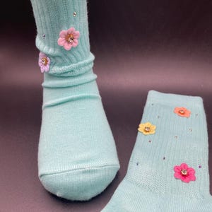 May include: A pair of light blue socks with colourful flower embellishments. The socks are ribbed and have a subtle sheen.