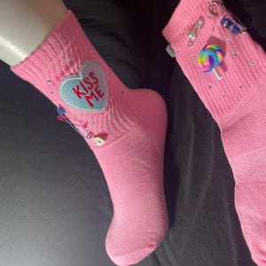May include: Pair of pink socks with embellishments. One sock features a heart-shaped patch with "KISS ME" text. Both socks have decorative pins, rhinestones, and a lollipop charm. The socks are displayed on a black surface.