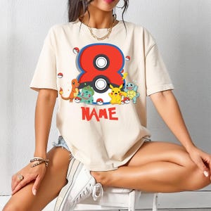 May include: Cream-colored t-shirt featuring a large red and blue number 8 design with a black and white center. The shirt includes cartoon characters and the word "NAME" in red. The design is centered on the front of the shirt.