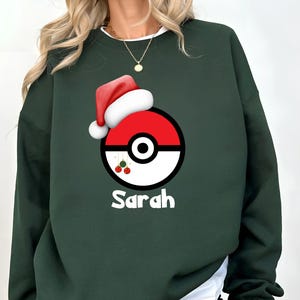 May include: Dark green sweatshirt featuring a red and white graphic resembling a Poké Ball, topped with a Santa hat. The name "Sarah" is printed in white below the graphic. The sweatshirt is a festive holiday design.