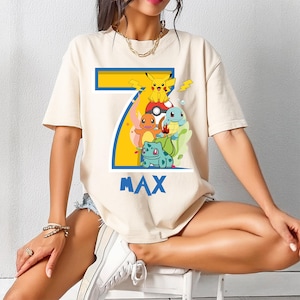 May include: A cream-colored t-shirt featuring a colorful graphic of the number 7 with various cartoon characters, including Pikachu. The word "MAX" is printed in blue below the graphic. The shirt has short sleeves.
