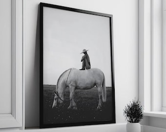 May include: Monochrome framed print showing a person seated on a white horse in a field. The horse is grazing, and the person is wearing a hat. The artwork is displayed on a white wall.