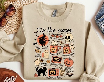 May include: Beige sweatshirt with a Halloween-themed graphic. The design includes a witch on a broom, pumpkins, a skull, and the text "'tis the season". Other elements include ghosts, bats, and the words "spooky witches" and "Halloween music".