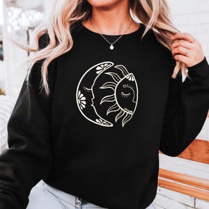 May include: Black sweatshirt featuring a white graphic of a crescent moon and sun with faces. The moon has floral accents, and the sun has closed eyes and rays. The design is centered on the chest.