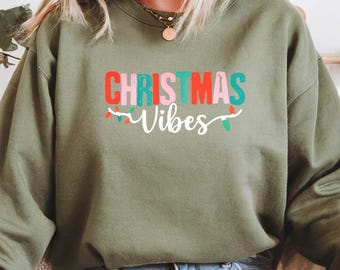 May include: An olive green sweatshirt with the words "CHRISTMAS Vibes" in colorful block letters and a string of lights design. The sweatshirt is a crew neck style and is made of a soft material.