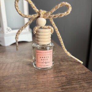 May include: A small glass bottle with a wooden cap and a natural twine bow. The bottle contains a natural oil diffuser labeled "Amber Noir oil diffuser 0.27 oz/8mL" and "Naturally Diffused".