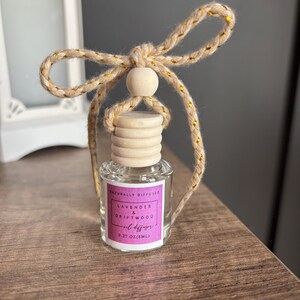 May include: A small glass bottle with a wooden cap and a gold and beige string tied around the top. The bottle contains a lavender and driftwood oil diffuser. The label reads "Naturally Diffused Lavender & Driftwood Oil Diffuser 0.27 oz (8mL)"