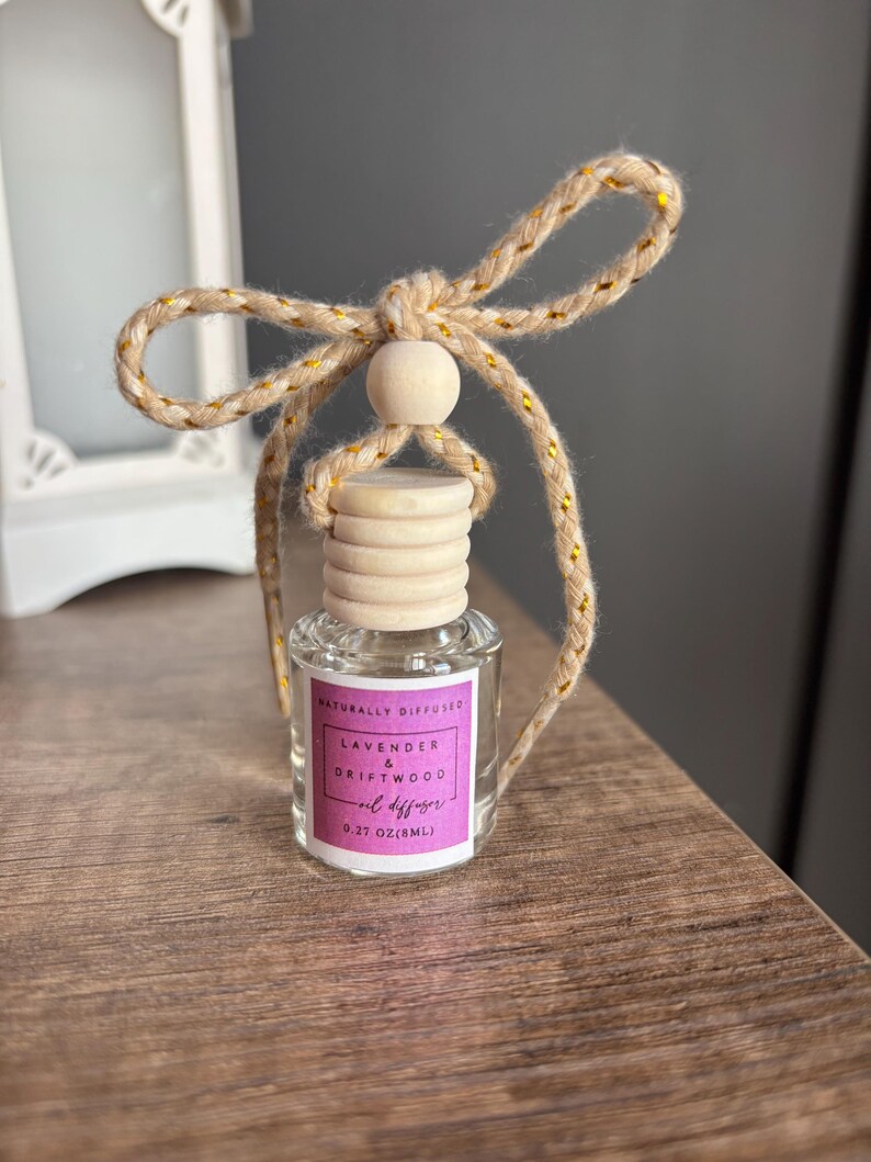May include: A small glass bottle with a wooden cap and a gold and beige string tied around the top. The bottle contains a lavender and driftwood oil diffuser. The label reads "Naturally Diffused Lavender & Driftwood Oil Diffuser 0.27 oz (8mL)"