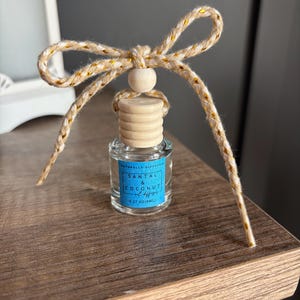 May include: A small glass bottle with a wooden cap and a gold and beige twine bow. The bottle contains a natural diffuser with the scent of Santal & Coconut, and the label reads "Naturally Diffused Santal & Coconut Oil Diffuser 0.37 oz (10mL)"