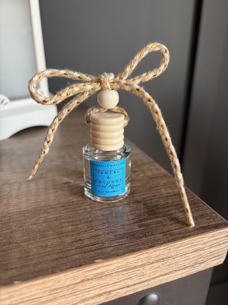 May include: A small glass bottle with a wooden cap and a gold and beige twine bow. The bottle contains a natural diffuser with the scent of Santal & Coconut, and the label reads "Naturally Diffused Santal & Coconut Oil Diffuser 0.37 oz (10mL)"