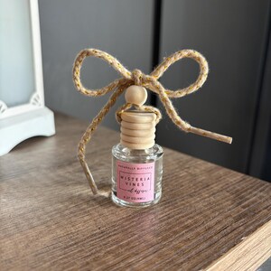 May include: A small glass bottle with a wooden cap and a gold string bow. The bottle is labeled "Naturally Diffused Wisteria Vines Oil Diffuser 0.27 oz (8ml)".