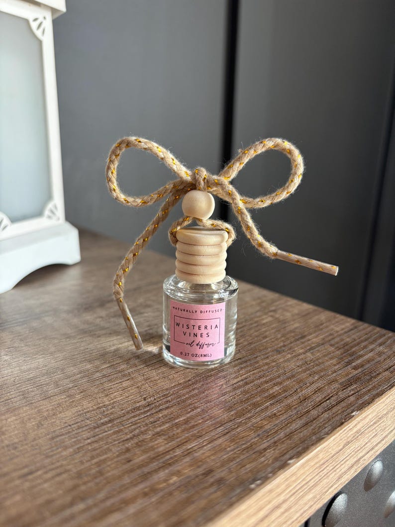May include: A small glass bottle with a wooden cap and a gold string bow. The bottle is labeled "Naturally Diffused Wisteria Vines Oil Diffuser 0.27 oz (8ml)".