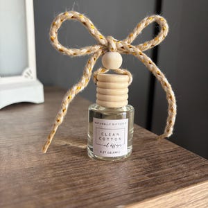 May include: A small glass bottle with a wooden cap and a brown and gold string tied around the top. The bottle contains a clear liquid and has a label that reads "Naturally Diffused Clean Cotton Oil Diffuser 0.27 oz (8mL)"