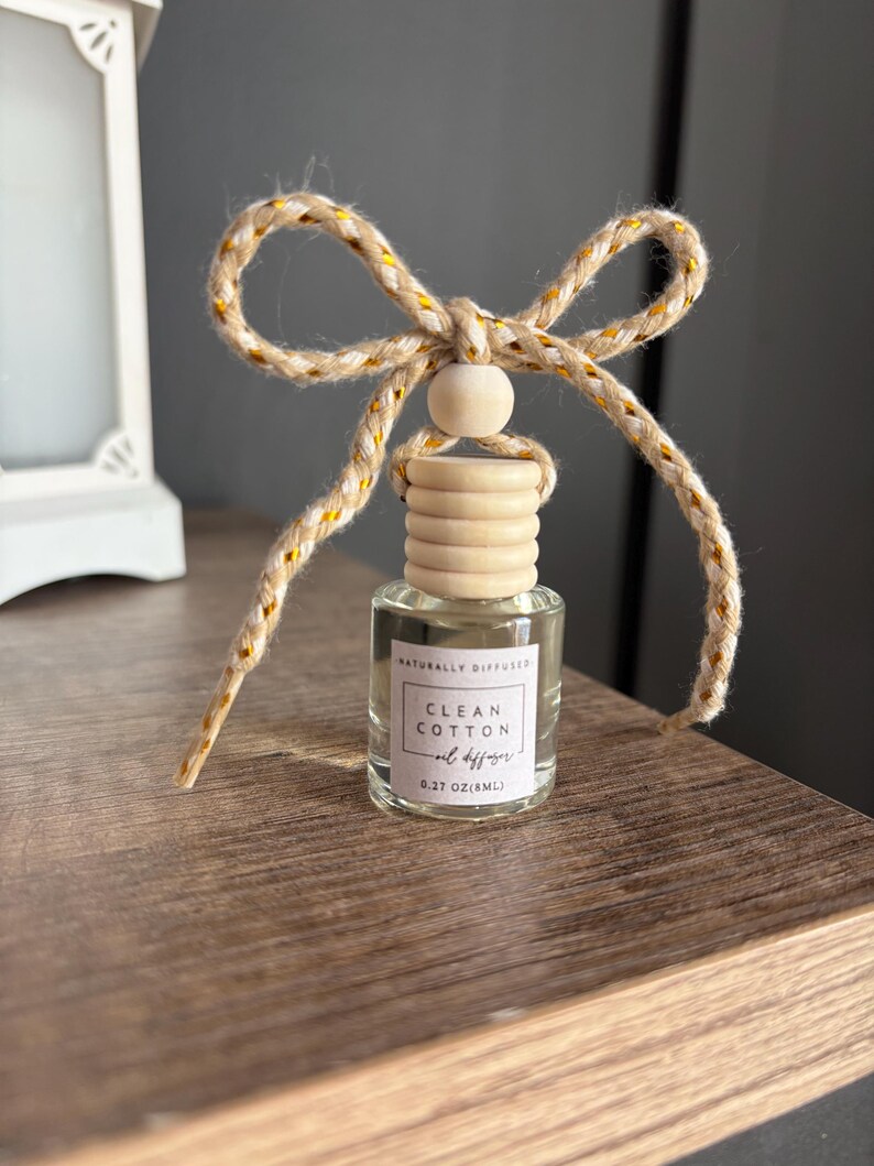 May include: A small glass bottle with a wooden cap and a brown and gold string tied around the top. The bottle contains a clear liquid and has a label that reads "Naturally Diffused Clean Cotton Oil Diffuser 0.27 oz (8mL)"