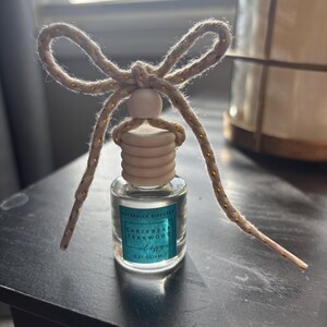 May include: A small glass bottle with a wooden cap and a gold string tied around it. The bottle contains a clear liquid and has a label that reads "Naturally Diffused Caribbean Teakwood Oil Diffuser 0.27 oz (8 ml)"