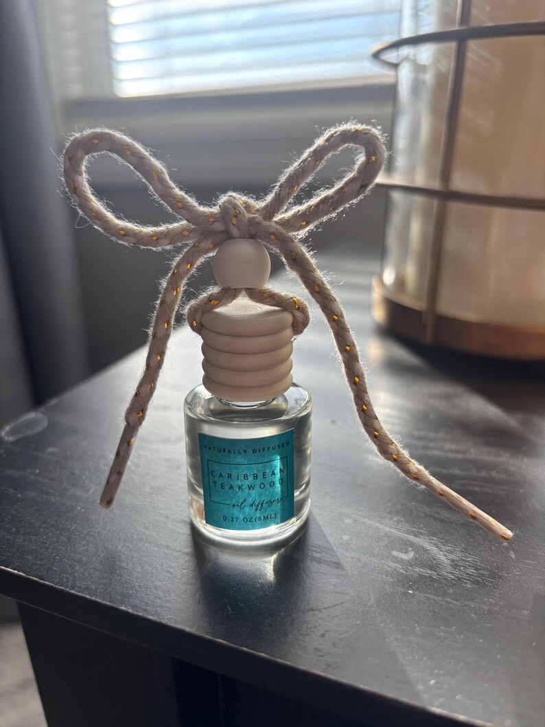 May include: A small glass bottle with a wooden cap and a gold string tied around it. The bottle contains a clear liquid and has a label that reads "Naturally Diffused Caribbean Teakwood Oil Diffuser 0.27 oz (8 ml)"