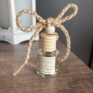 May include: A small glass bottle with a wooden cap and a gold and white string tied around it. The bottle contains a clear liquid and has a label that reads "Naturally Diffused Petals & Tangerine Oil Diffuser 0.27 oz (8ml)"