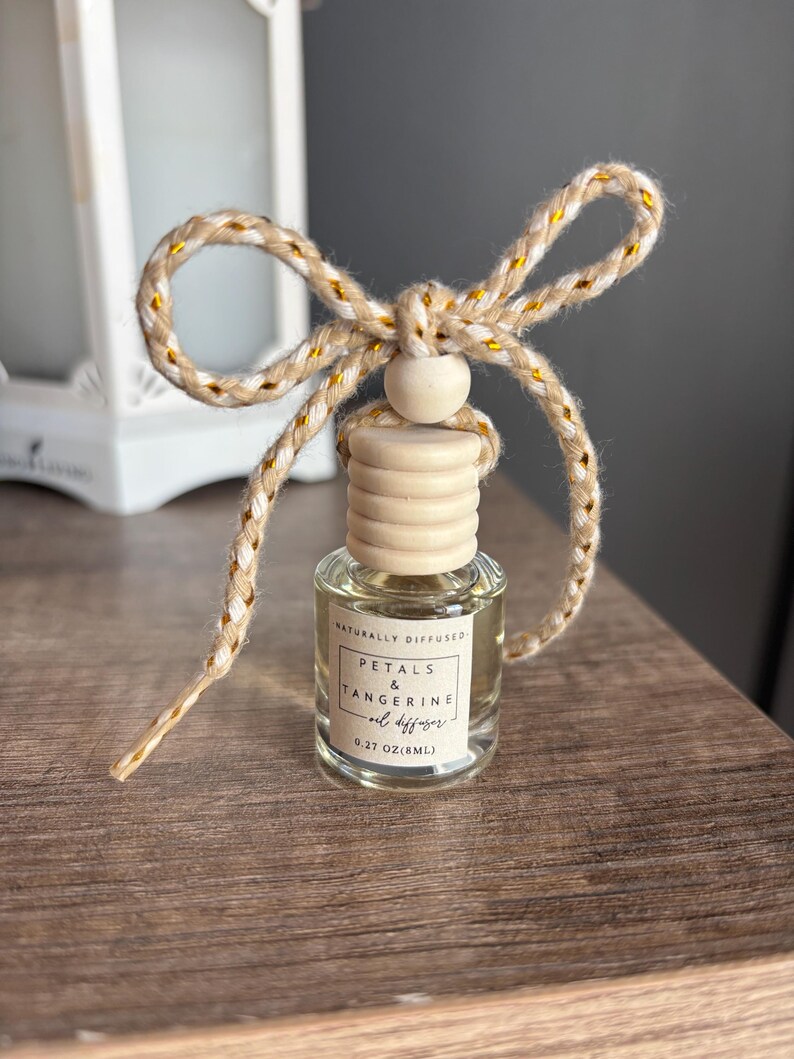May include: A small glass bottle with a wooden cap and a gold and white string tied around it. The bottle contains a clear liquid and has a label that reads "Naturally Diffused Petals & Tangerine Oil Diffuser 0.27 oz (8ml)"