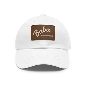 May include: White baseball cap with a curved brim. A brown rectangular patch on the front displays the word "Baba" in a cursive font above "Komandant". The cap is made of a soft, breathable fabric.