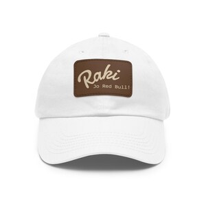 Op de afbeelding: Witte baseballcap met een gebogen klep. De cap heeft een bruin rechthoekig embleem met het woord "Raki" in een cursief lettertype en de tekst "Jo Red Bull!" eronder. De cap is gemaakt van een zacht materiaal.