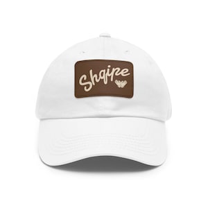 Op de afbeelding: Witte baseballpet met een gebogen klep. Een bruin rechthoekig label op de voorkant toont het woord "Shaire" in cursief schrift, met een kleine afbeelding van twee handen eronder. De pet is gemaakt van een zachte stof.