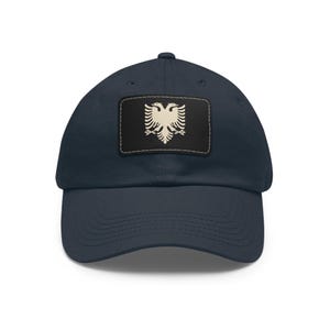 May include: Navy blue baseball cap with a rectangular patch featuring a cream-colored double-headed eagle emblem on a black background. The cap has a curved brim and a classic six-panel design.
